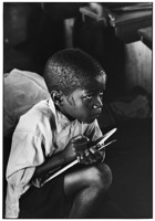 Earnest boy squats on haunches and strains to follow lesson in heat of packed classroom.
